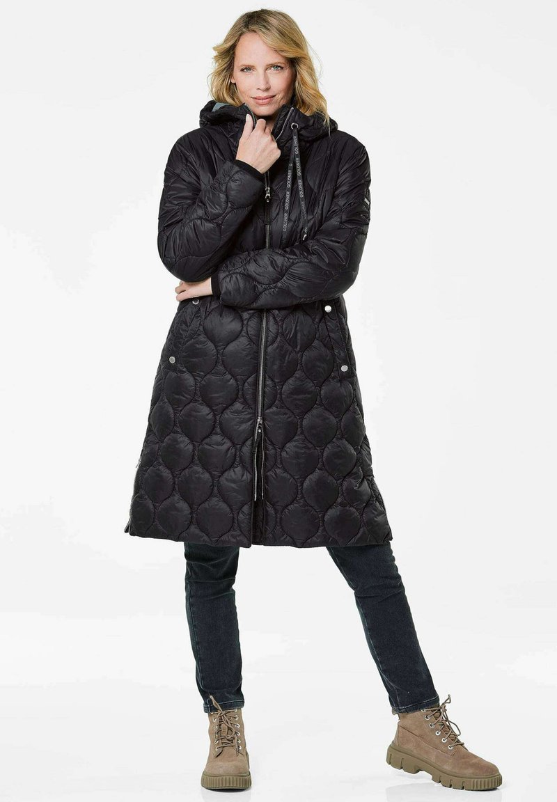 Zwarte gewatteerde pufferjas met een capuchon, voorzien van een rits aan de voorkant en een trekkoord, gecombineerd met donkere jeans en tan kleurige veterschoenen.