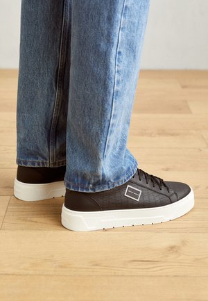 Zapatillas negras con material texturizado, que presentan una suela y cordones blancos, combinadas con jeans de mezclilla azul claro. Fondo de suelo de madera.