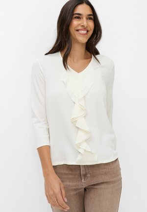 Vrouw met lang donker haar glimlacht, draagt een witte blouse met ruches aan de voorkant en driekwartmouwen, gecombineerd met lichtbruine broek.