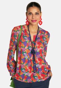 A woman with long hair wears a vibrant floral blouse, red heart earrings, and a green bag, smiling against a neutral background.