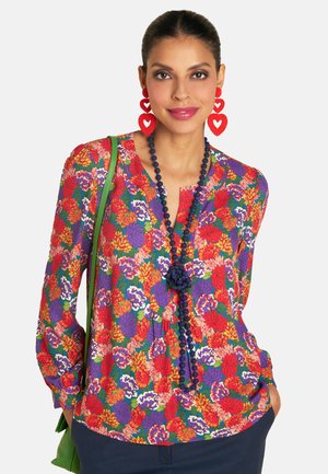 Eine Frau mit langen Haaren trägt eine lebhafte Blumenbluse, rote Herzohrringe und eine grüne Tasche und lächelt vor einem neutralen Hintergrund.