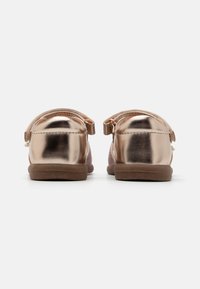 Zapatos metálicos color rosa dorado con una textura suave, puntera redondeada y una suela plana marrón. Cuentan con una correa en la parte posterior para facilitar el uso.