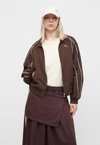 Γυναίκα που φοράει καφέ μπουφάν Puma, εκρού πουκάμισο, σκούρα καφέ πολυεπίπεδη φούστα και λευκό καπέλο Puma, στέκεται μπροστά σε απλό ανοιχτό φόντο.