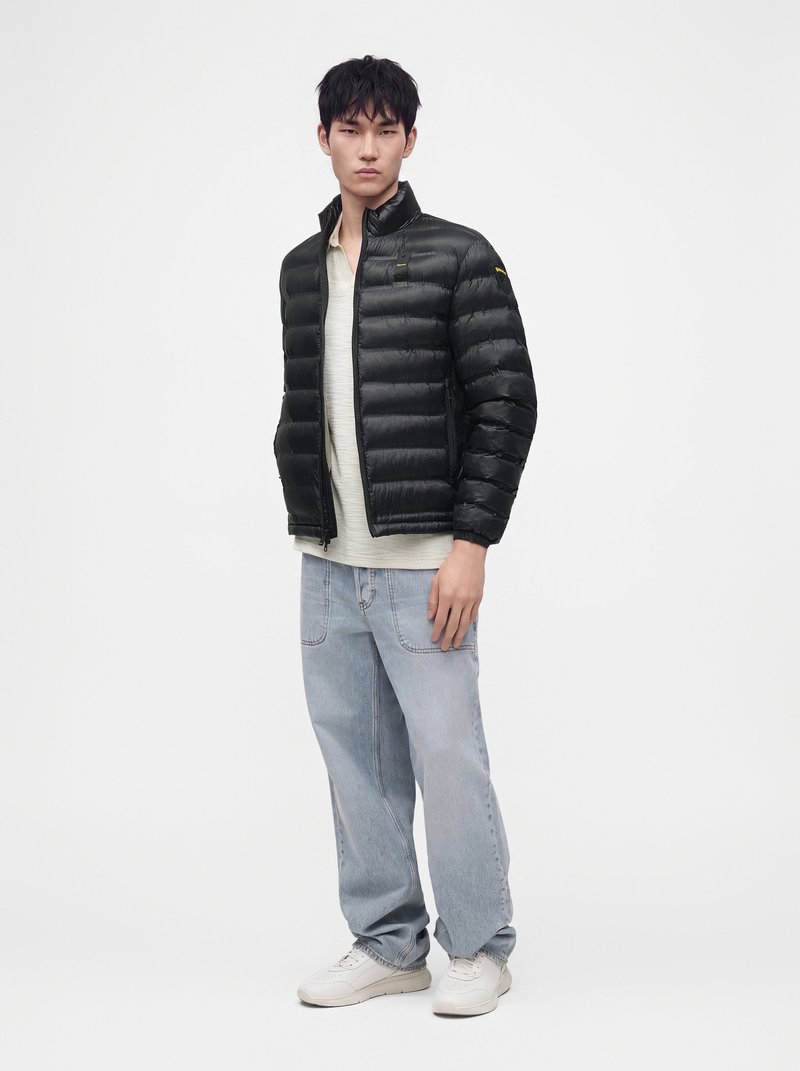 Jeune homme portant une doudoune noire, une chemise beige clair, un jean bleu clair et des baskets blanches, debout devant un fond blanc.
