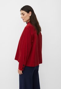 Červený kordurový jacket s vysokým límcem a dlouhými rukávy, se třemi černými knoflíky na každém manžetě. Nosí se přes tmavě modré kalhoty s širokými nohavicemi.