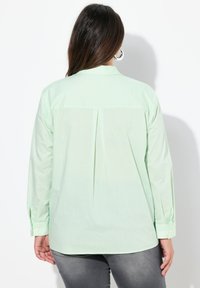 Camicia a maniche lunghe di colore verde chiaro, realizzata in tessuto morbido con strisce verticali bianche, dotata di una piega sul retro e polsini con bottoni.