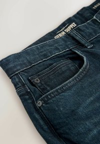 Jeans din denim închis, cu o textură netedă, prezentând cusături intricate, un detaliu de buzunar și un etichetă pe talie cu text alb.
