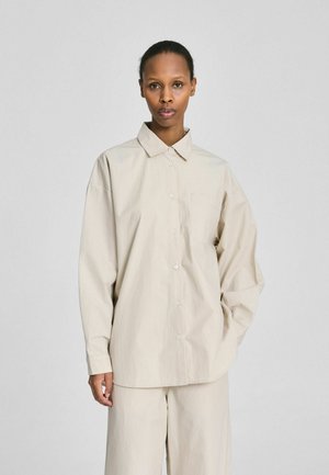 Vrouw in een loszittend beige overhemd met knopen en bijpassende wijd uitlopende broek, staand tegen een effen lichtgrijze achtergrond.