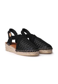 Espadrille negra tejida con una suela gruesa de esparto, talón abierto y tiras elásticas entrecruzadas. Interior de cuero suave con un acabado natural.