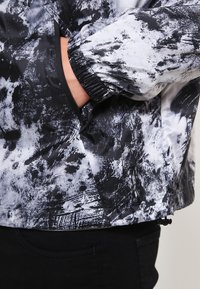 Lekka kurtka z abstrakcyjnym czarno-białym nadrukiem, z elastycznymi mankietami i bocznymi kieszeniami, o błyszczącej, gładkiej fakturze i nowoczesnym designie.