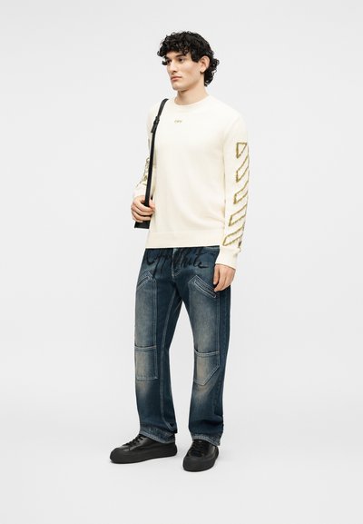 Άνδρας που φορά λευκό πουλόβερ με διαγώνια σχέδια στα μανίκια, τζιν με μπλε patches, μαύρα παπούτσια και κρατά μαύρη τσάντα ώμου.