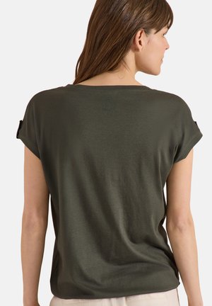 Kobieta w ciemnozielonym krótkim rękawem z podwiniętymi mankietami i subtelnym okrągłym logo na górnej części pleców, zwrócona tyłem na jednolitym tle.
