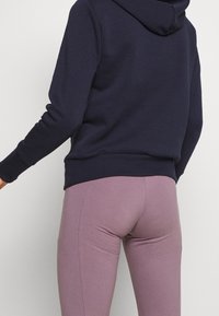 Granatowa bluza z kapturem i długimi rękawami w połączeniu z dopasowanymi fioletowymi legginsami, prezentującymi gładką teksturę i bezszwowy design.