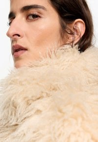Abrigo de piel sintética en color crema, con una superficie suave, texturizada y desaliñada. Primer plano del rostro que muestra un maquillaje natural y joyas en las orejas.