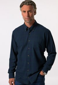 Camisa a cuadros en tonos azul marino, fabricada con un tejido suave con líneas de rejilla verdes, con cuello button-down, bolsillo en el pecho y botones marrones, llevada sin meter por dentro con unos jeans azules.