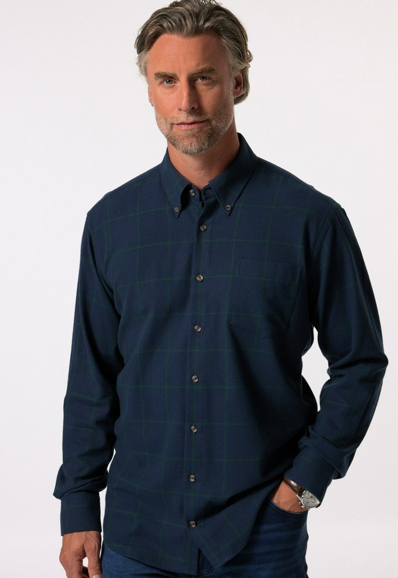 Camisa a cuadros en tonos azul marino, fabricada con un tejido suave con líneas de rejilla verdes, con cuello button-down, bolsillo en el pecho y botones marrones, llevada sin meter por dentro con unos jeans azules.