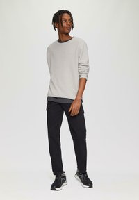 Hellgrauer, strukturierter Pullover mit rundem Ausschnitt, kombiniert mit schwarzen Cargo-Hosen mit Seitentaschen und schwarzen Sneakers mit weißen Akzenten.