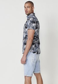 Camicia a maniche corte con stampa tropicale in bianco e nero, abbinata a pantaloni shorts in denim azzurro chiaro arrotolati, con tasche posteriori e dettagli dorati.
