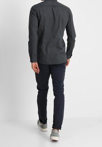 Donkergrijze blouse met lange mouwen en een subtiel patroon, gecombineerd met marineblauwe broek en grijze sneakers. Heeft een kraag en knoop manchet.