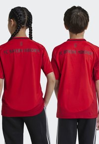 Maillot de football rouge du FC Bayern München, manches courtes, col rond. Texte noir au dos avec "FC BAYERN MÜNCHEN". Tissu lisse, coupe décontractée.