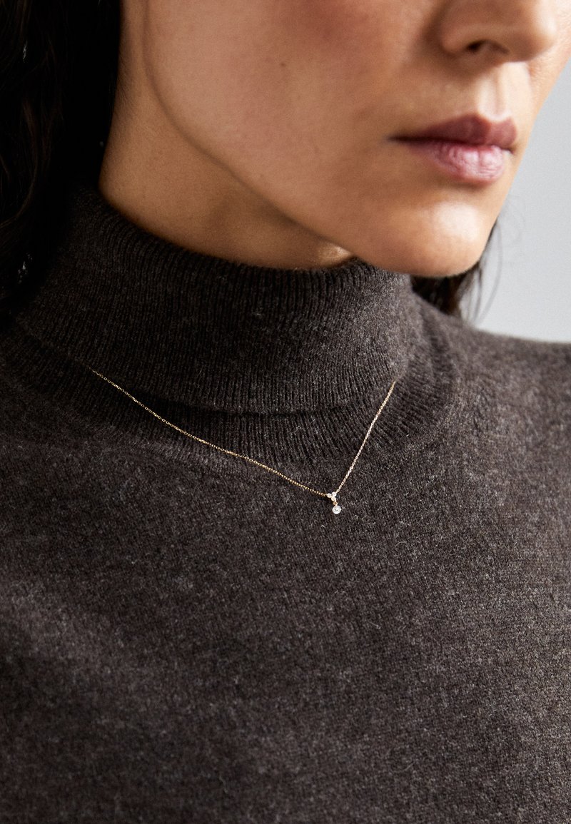 Maglione a collo alto in maglia marrone con una trama liscia, abbinato a una delicata collana d'oro con un piccolo pendente rotondo.