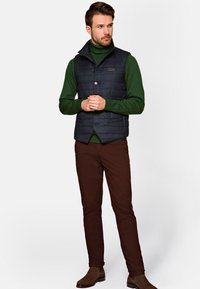 Granatowa pikowana kamizelka z kołnierzem stójkowym, zapięciem na guziki, noszona na zielonym golfie, w połączeniu z brązowymi spodniami i brązowymi butami.