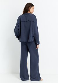 Diep blauw, textiel outfit met een losse blouse met rafelige randen en wijde broekspijpen. Knopdetail op de mouwen.