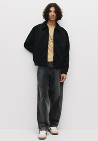 Giacca bomber nera in suede, t-shirt grafica gialla, jeans a gamba larga grigio scuro e sneakers bianche con dettagli marroni. Design minimalista.
