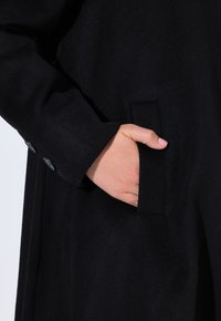 Abrigo de lana negra con un bolsillo lateral de textura suave. Los detalles de los botones son visibles en el puño de la manga.