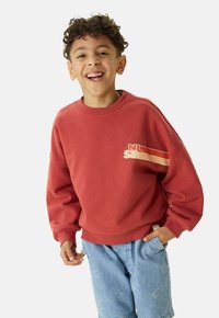 Un sweat-shirt rouge à col rond et à coupe oversize, orné des initiales "NL" et "SB" dans des tons neutres. Associé à un short bleu clair, décoré d'un motif de smileys.