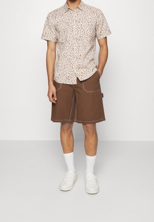 Kortärmad skjorta med ett vitt och brunt blommönster, kombinerad med bruna shorts med vit sömnad. Bärs med vita strumpor och skor.