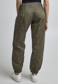 Pantalon léger vert olive avec une taille élastique, des poignets froncés et une petite étiquette logo à l'arrière, associé à des baskets blanches.