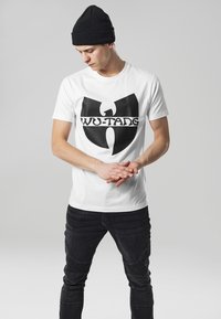 Balta medvilninė marškinė su dideliu juodu Wu-Tang logotipu, derinami su juodomis džinsų kelnėmis. Modelis vilki juodą megztinį, stovėdamas su sukryžiuotomis rankomis.