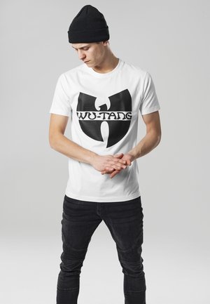 Biely bavlnený tričko s veľkým čiernym logom Wu-Tang, v kombinácii s čiernymi džínsami. Model má na hlave čiernu čiapku a stojí s prekríženými rukami.