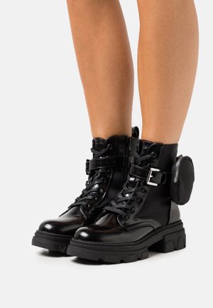 Schwarze Lackleder-Stiefeletten mit einer klobigen Sohle, Schnürdesign, seitlichem Reißverschluss, Schnallen-Detail und einem kleinen Täschchen auf der Rückseite.