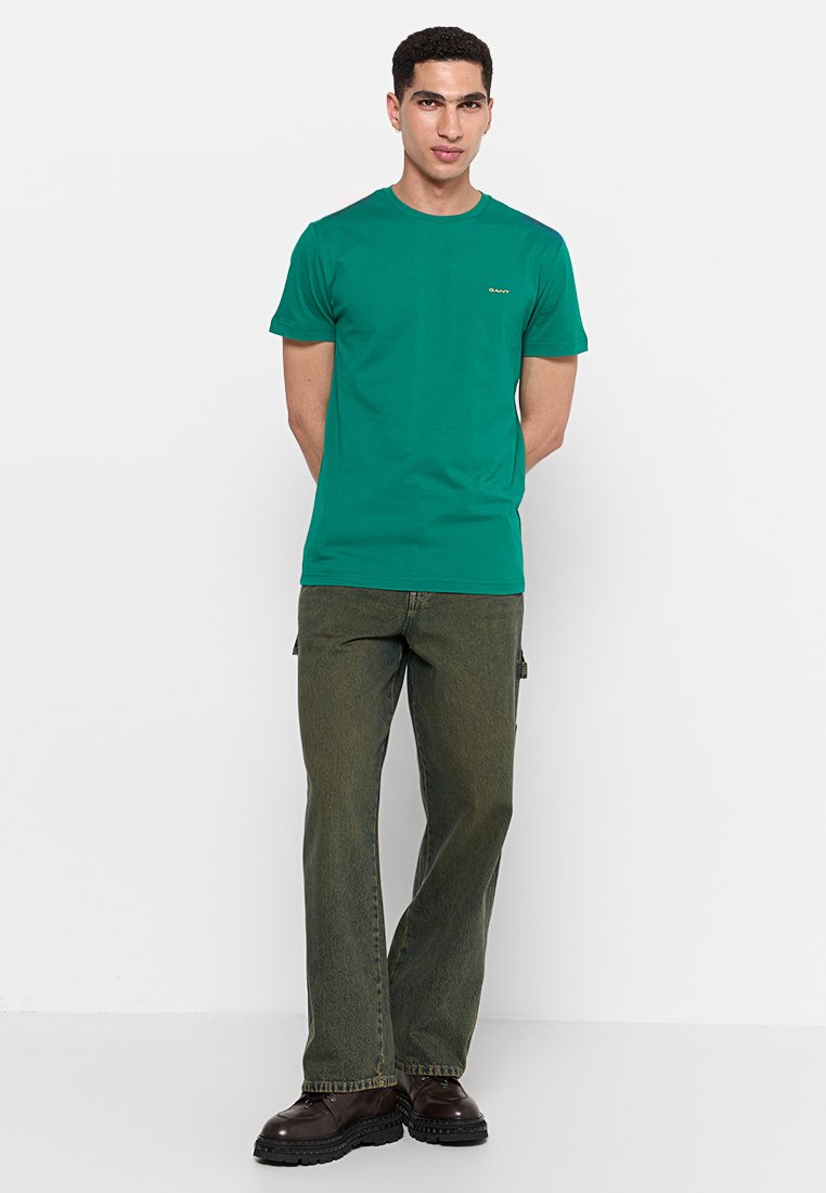 Camiseta de algodón verde con mangas cortas y cuello redondo, que presenta un pequeño logo en el pecho, combinada con pantalones cargo de color verde oliva.