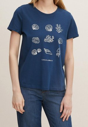Kvinde iført en mørkeblå t-shirt med hvide havskaller og koraltrykker og teksten "Life is a beach" kombineret med blå jeans.