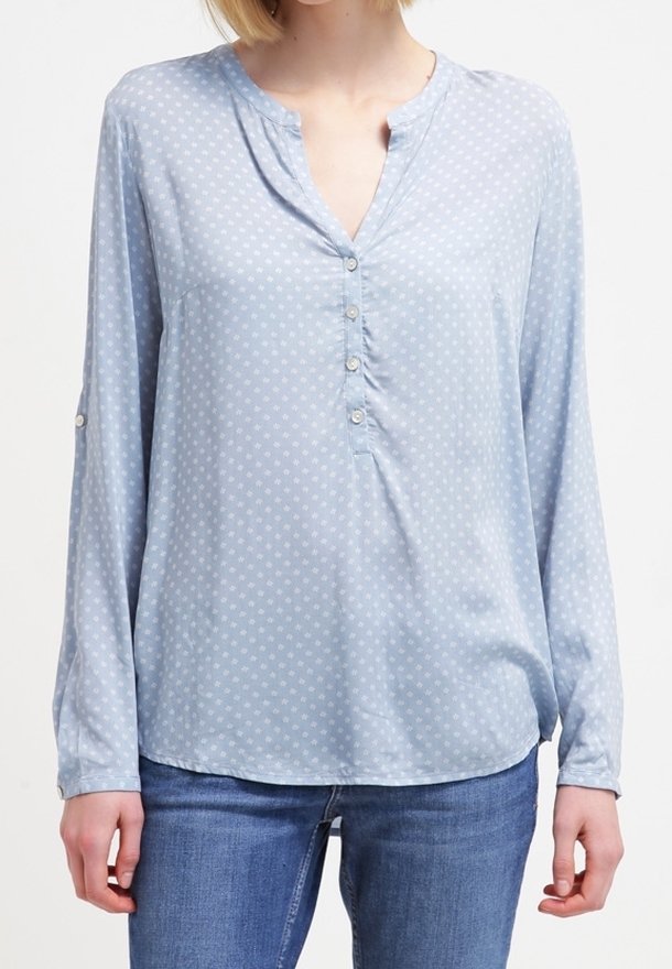 Blusa azzurro chiaro con scollatura a V, maniche lunghe e pois bianchi. Presenta una pattina con bottoni e un orlo curvato.