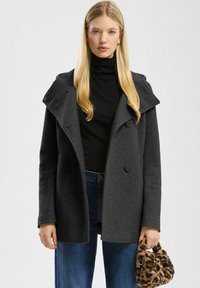 Zwarte, gestructureerde jas met een brede kraag, dubbele knoopsluiting en lange mouwen. Gecombineerd met donkere jeans en een handtas met luipaardprint.