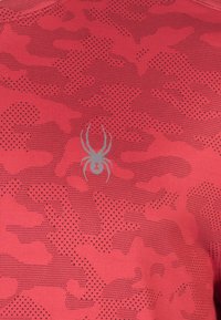Rød atletisk skjorte med et struktureret camouflage-mønster og et gråt edderkop-logo i midten. Fremstillet af letvægts, åndbart materiale.