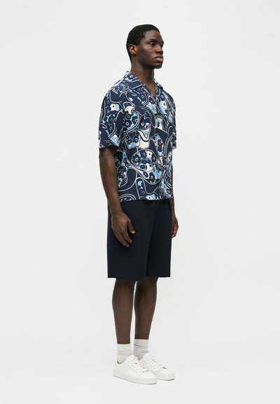 Άνδρας που φοράει κοντομάνικο μπλε πουκάμισο με σχέδιο, ναυτικό σορτς, λευκές κάλτσες και λευκά αθλητικά παπούτσια στέκεται μπροστά σε μια απλή φόντο.