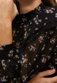 Blusa nera a fiori con tessuto trasparente, collo alto e dettagli arricciati. Delicati fiori rosa e foglie verdi in tutto il design.