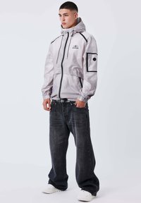 Giovane uomo con giacca a vento grigia chiara, jeans neri larghi e sneakers bianche, in piedi con le mani in tasca su uno sfondo semplice.