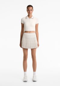 Bílé krátké polo tričko s zipovým límcem a krátkými rukávy, doplněné o odpovídající skládací sukni. Tenisky dolaďují outfit.