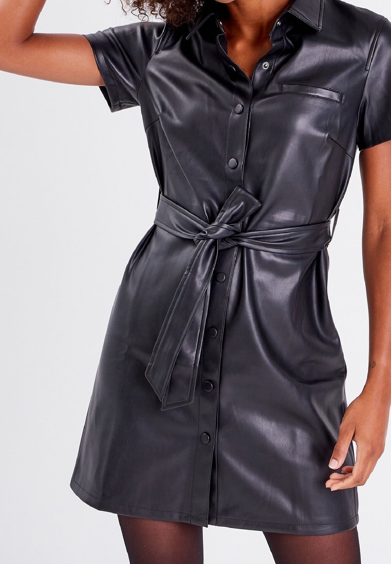 Kvinna som bär en svart kortärmad klänning i konstläder med knappar och ett knytbälte i midjan.