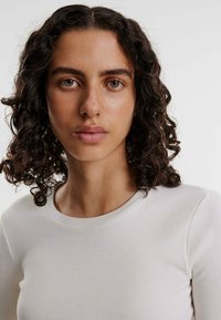 Nahaufnahme einer Person mit lockigem, dunklem Haar, die ein hellbeiges, geripptes Langarmoberteil mit rundem Ausschnitt trägt. Glatte Haut und neutraler Hintergrund.