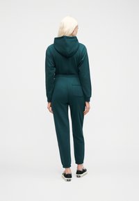 Γυναίκα με ξανθά μαλλιά που φοράει πετρόλ φούτερ με κουκούλα και αντίστοιχο παντελόνι φόρμας με πίσω τσέπη, όρθια με την πλάτη προς τα εμπρός σε λευκό φόντο.