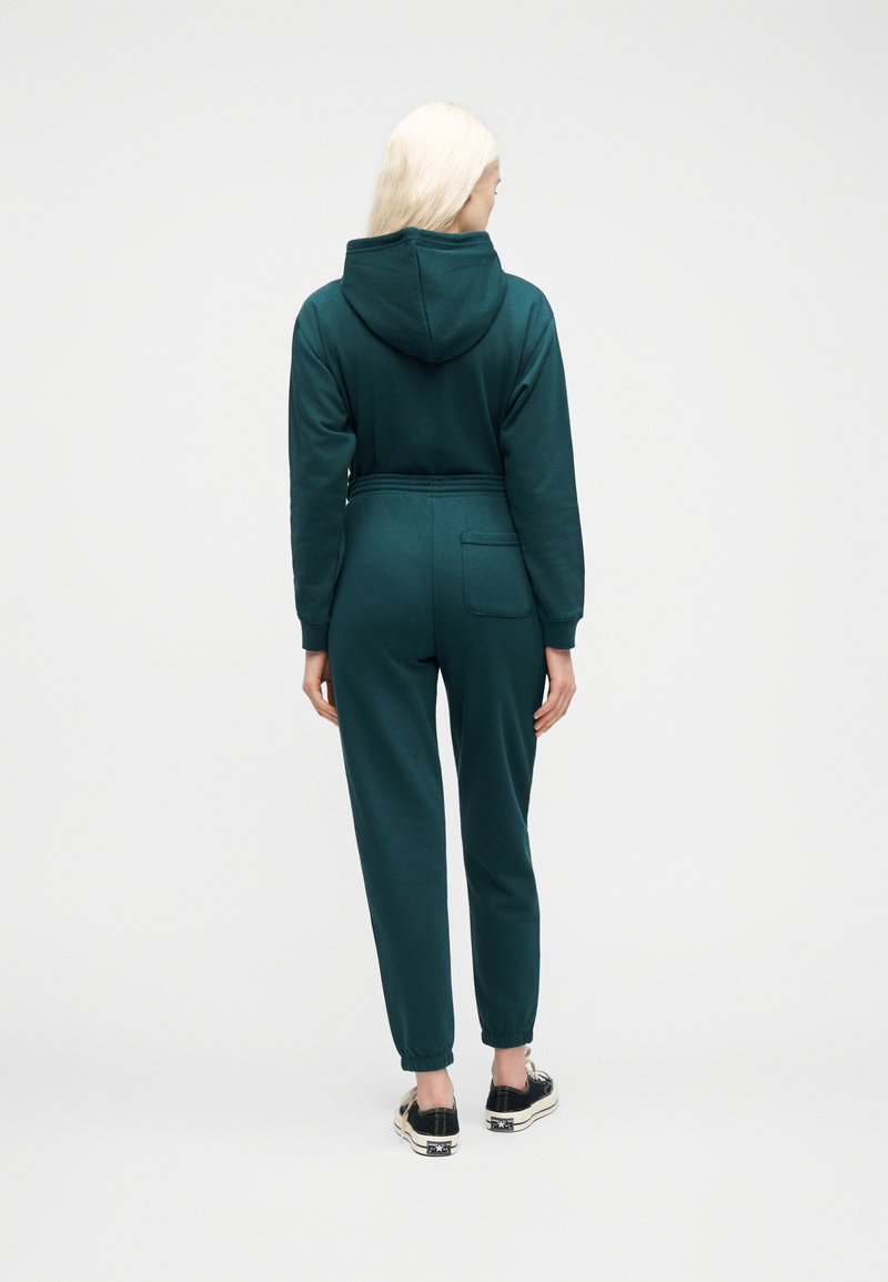 Woman with blonde hair wearing a teal hooded sweatshirt and matching jogger pants with back pocket, standing facing away on a white background.