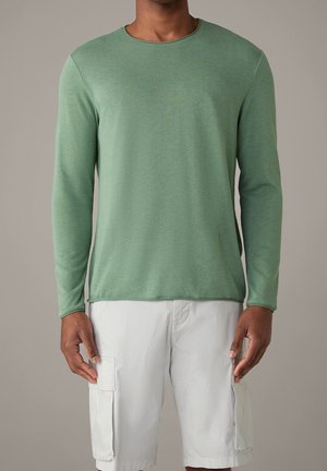 Homme portant une chemise à manches longues vert clair unie et un short cargo blanc, debout devant un fond neutre.