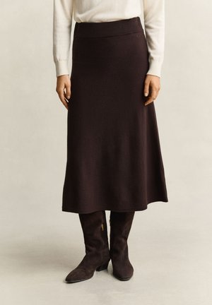 Frau trägt einen dunkelbraunen Midi-Rock, cremefarbenes Langarmshirt und dunkelbraune Wildlederstiefel bis zur Mitte der Waden, steht vor einem einfarbigen Hintergrund.